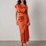 Woman in an orange satin wrap midi dress with a draped bodice and tied waist, holding a beige clutch.