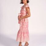 Woman modeling a pink lace midi dress with a tied waist, scalloped edges, pearl necklace, and strappy heels in a studio.