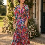 Woman in a colorful floral dress stands on a gravel path beside a stone house with roses, smiling.
