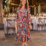 Smiling woman in a bright floral midi dress with a coral fascinator and beige clutch at a rustic reception venue.