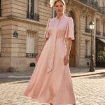 Woman in a light pink midi dress with a long scarf-like tie, wearing a large white fascinator, standing on a Parisian street.
