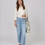 Woman wearing a cream button-down blouse and light blue wide-leg jeans, carrying a black-and-white patterned tote bag, wearing brown slide sandals.
