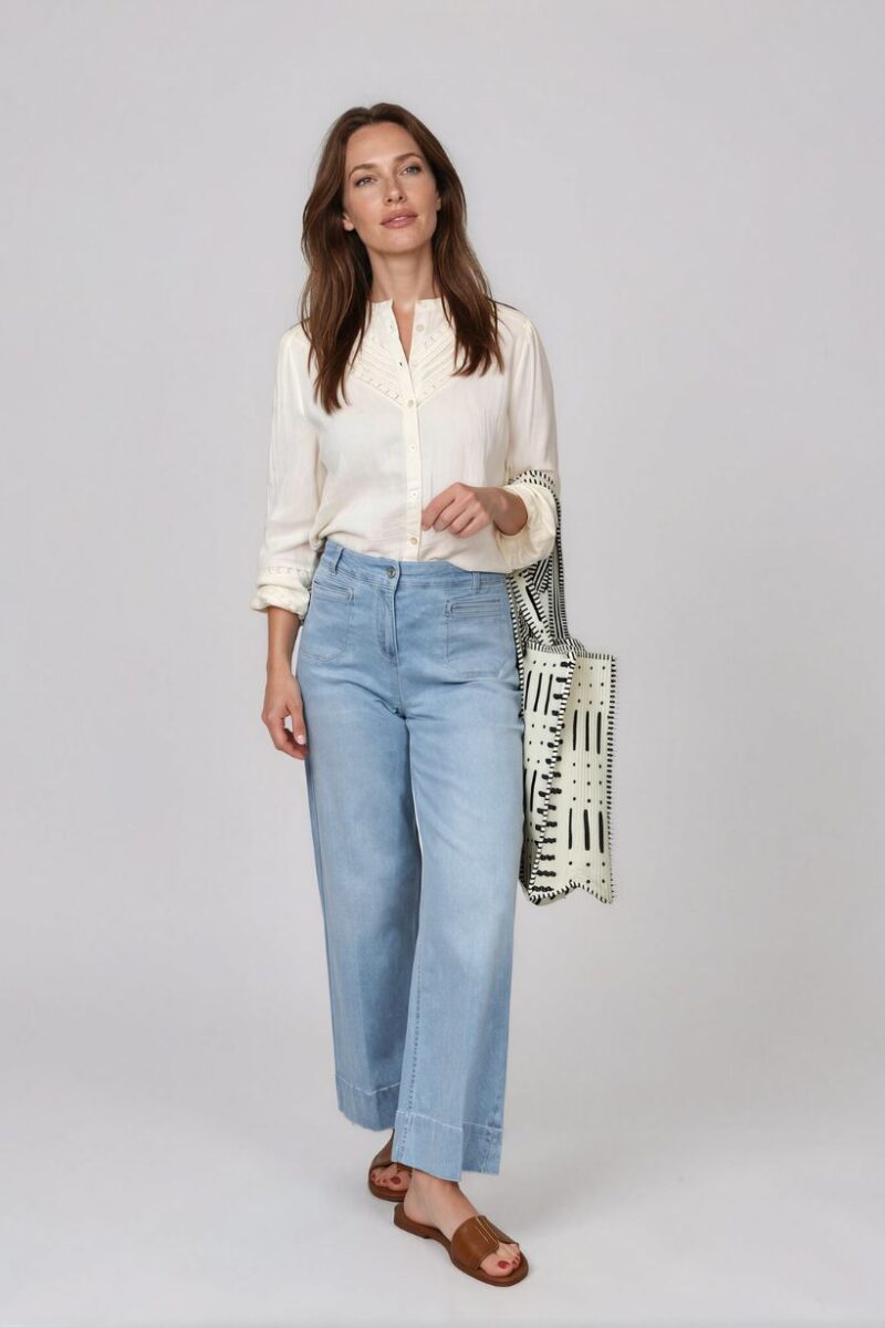 Woman wearing a cream button-down blouse and light blue wide-leg jeans, carrying a black-and-white patterned tote bag, wearing brown slide sandals.