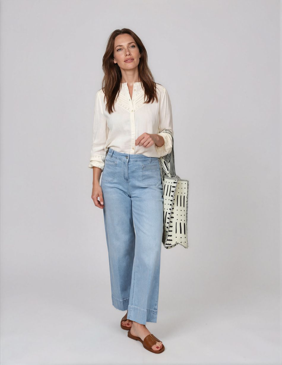 Robell-Debby-light-denim Woman wearing a cream button-down blouse and light blue wide-leg jeans, carrying a black-and-white patterned tote bag, wearing brown slide sandals.