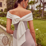 Back view of a woman in an off‑the‑shoulder lavender gown with a large white bow tied at the back, in a garden setting.