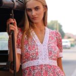Young woman with blonde hair leaning against a vehicle door, wearing a pink floral dress with white lace trim in a street setting.