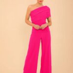 Woman wearing a hot-pink one-shoulder jumpsuit with wide-leg pants, standing against a beige backdrop, hands clasped in front.