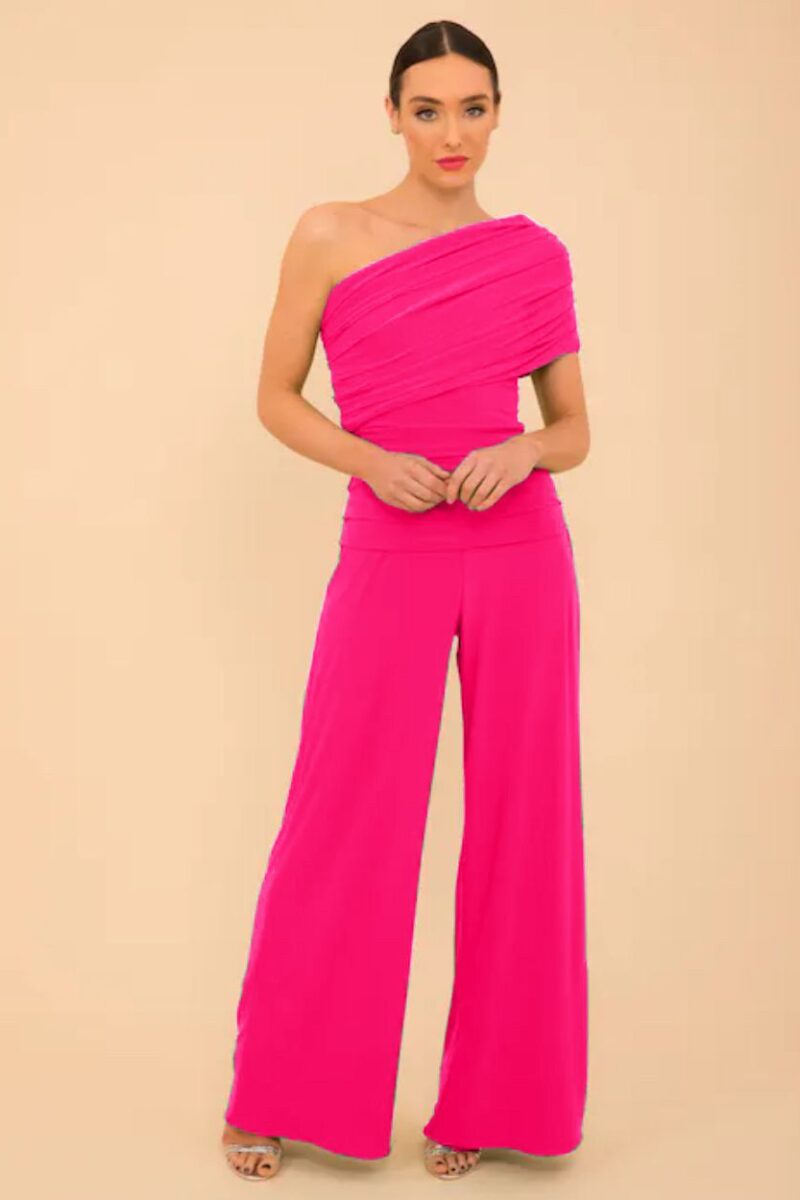 Woman wearing a hot-pink one-shoulder jumpsuit with wide-leg pants, standing against a beige backdrop, hands clasped in front.