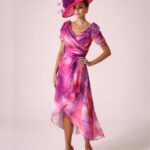 Model in a pink and purple tie-dye wrap dress with a matching wide-brim pink hat and pink heels, posing with hand on hip.