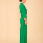 Side profile of a woman wearing a bright green, full-length outfit with long sleeves, standing against a beige background.