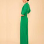 Profile view of a woman with dark hair in a bright green, full-length jumpsuit posing with hands on hips against a beige background