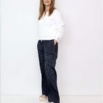 Woman standing against a white wall in a white cardigan, navy pinstripe wide-leg pants, and gold shoes; hands in pockets, looking at camera.