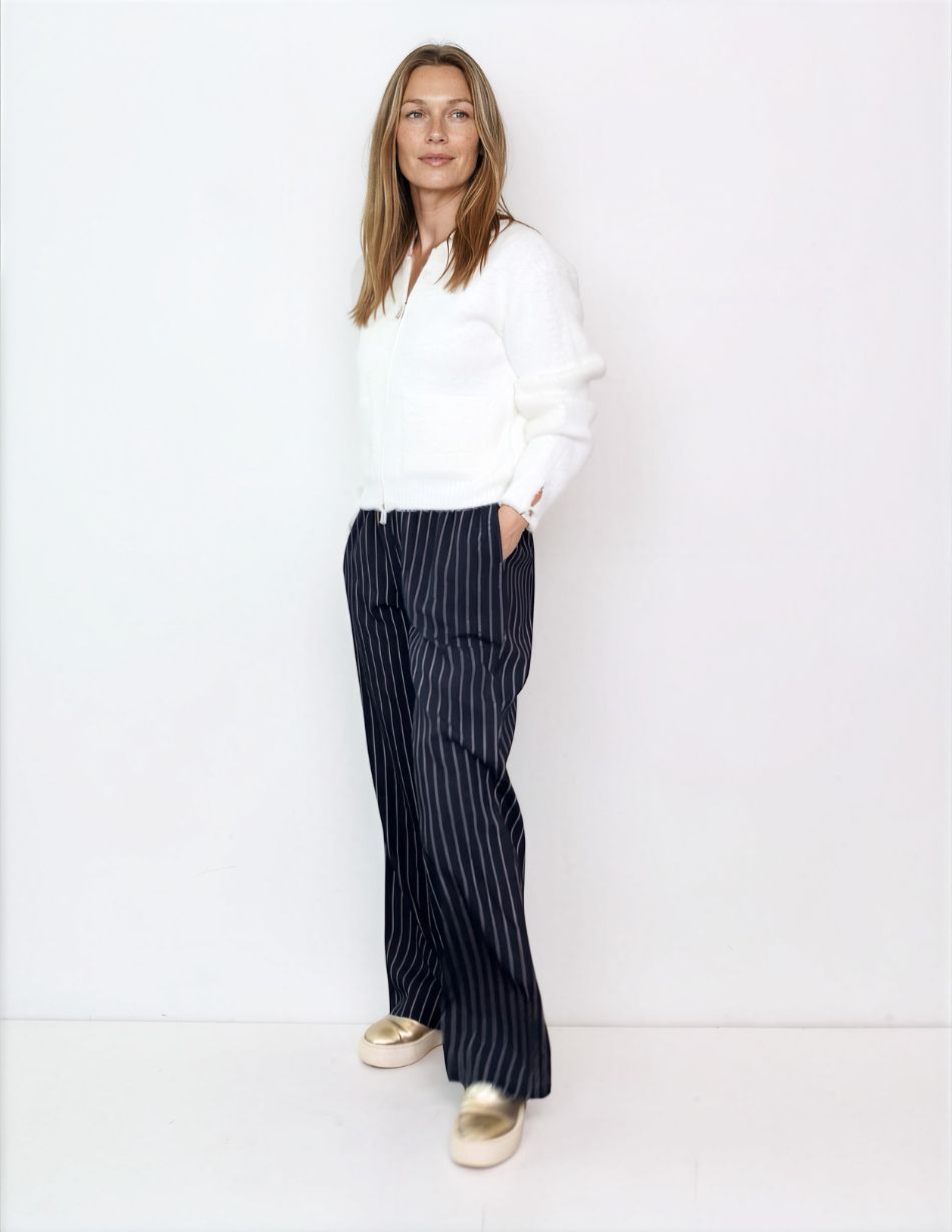 Woman standing against a white wall in a white cardigan, navy pinstripe wide-leg pants, and gold shoes; hands in pockets, looking at camera.