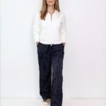 Woman stands facing camera in a white zip sweater and navy pinstripe wide-leg pants, with gold shoes, against a white background.