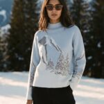 Woman standing in a snowy landscape wearing a light blue sweater with a mountain print and dark pants, sunglasses.