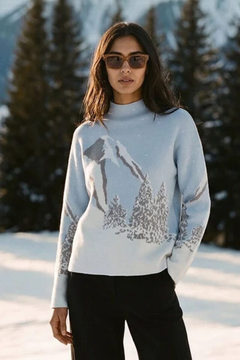 Woman standing in a snowy landscape wearing a light blue sweater with a mountain print and dark pants, sunglasses.