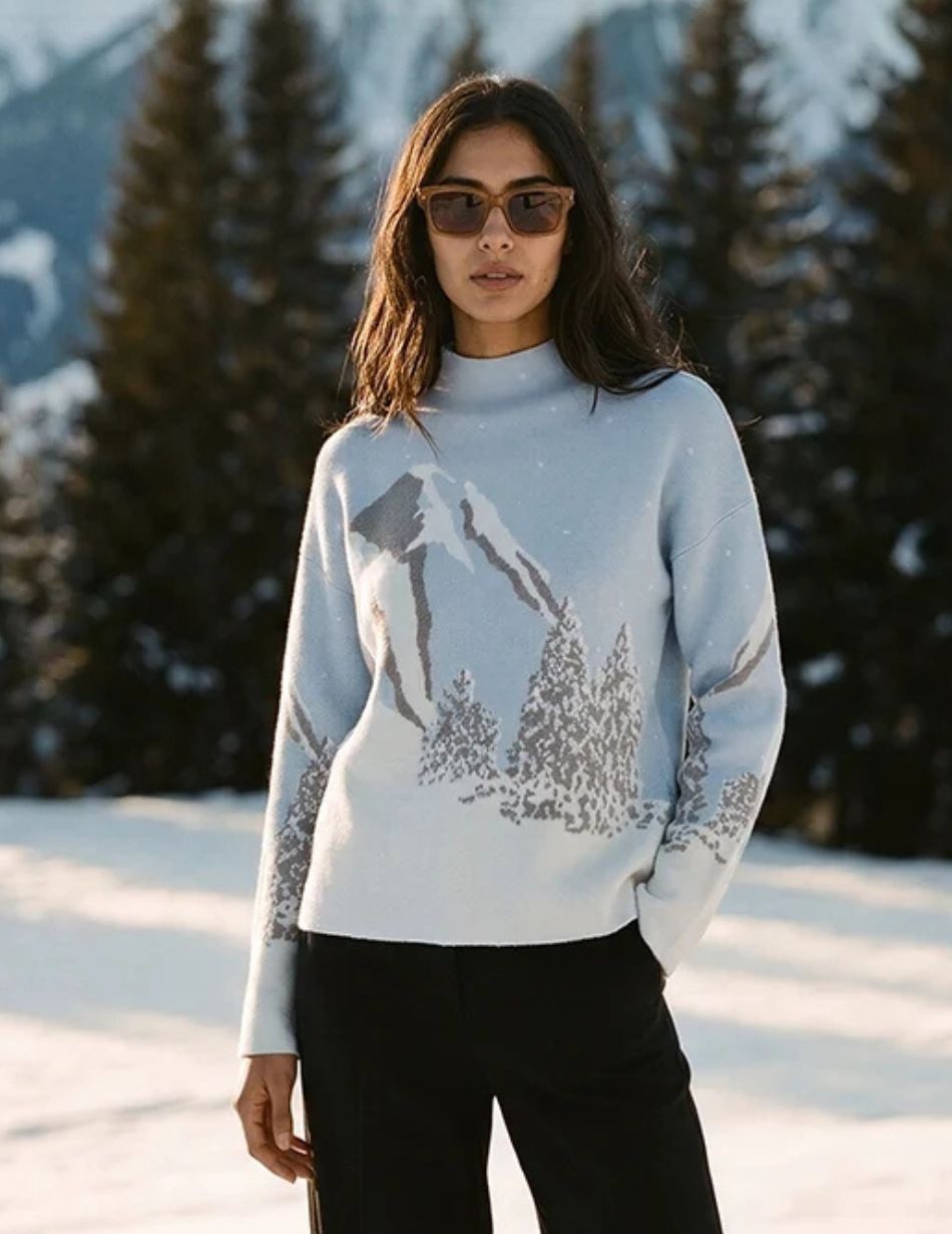 Alison Sheri Ski Scene Jumper A48302 Woman standing in a snowy landscape wearing a light blue sweater with a mountain print and dark pants, sunglasses.