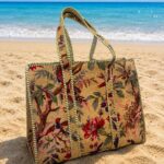 Woven floral beach bag on sand with blue ocean in the background, capturing a sunny seaside scene.