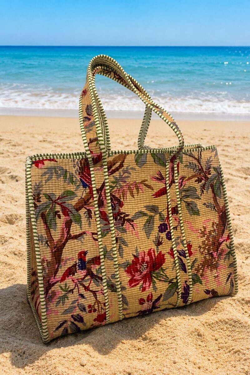 Woven floral beach bag on sand with blue ocean in the background, capturing a sunny seaside scene.