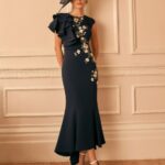Woman in a navy midi dress with floral embroidery and a dramatic one-shoulder ruffle, wearing a black fascinator, standing in a beige paneled room.