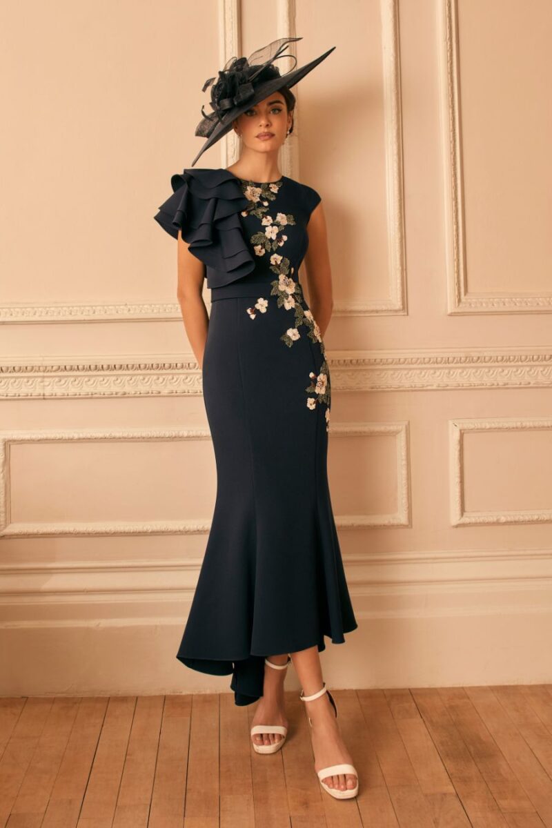 Woman in a navy midi dress with floral embroidery and a dramatic one-shoulder ruffle, wearing a black fascinator, standing in a beige paneled room.