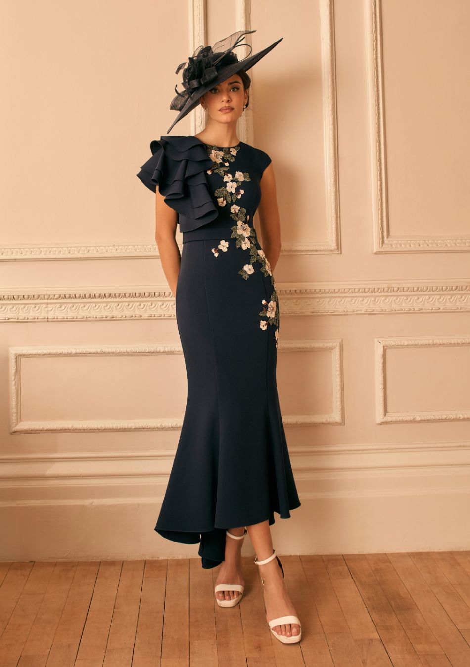 Woman in a navy midi dress with floral embroidery and a dramatic one-shoulder ruffle, wearing a black fascinator, standing in a beige paneled room.