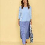 Woman standing against a pale yellow wall in a light blue long-sleeve top and a blue patterned ankle-length skirt, wearing tan sandals and carrying striped bags.