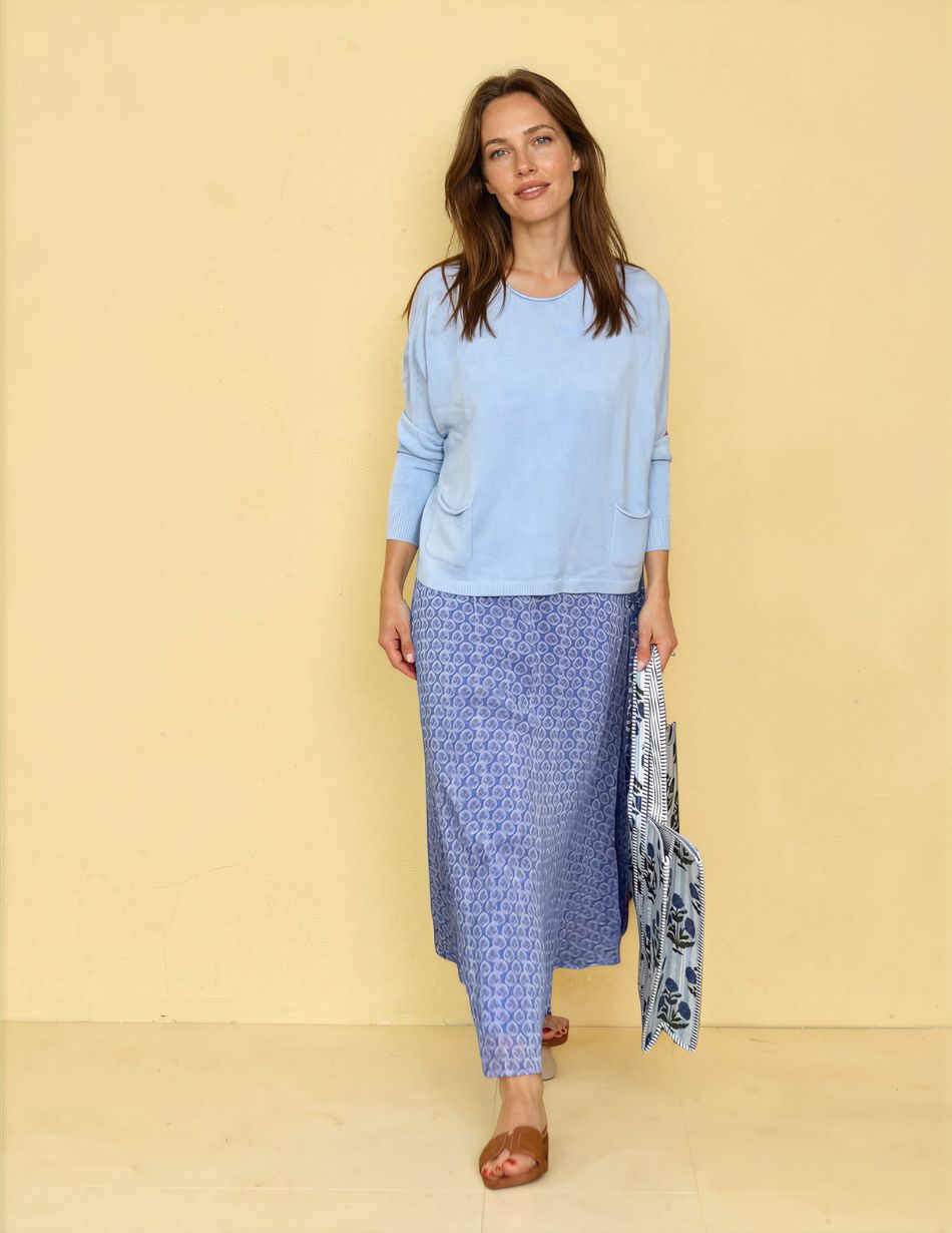 Woman standing against a pale yellow wall in a light blue long-sleeve top and a blue patterned ankle-length skirt, wearing tan sandals and carrying striped bags.