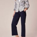 Woman modeling an outfit: snakeskin-pattern blazer over a white top, navy ankle-length pants, and white slip-on sneakers, with one hand in her pocket.