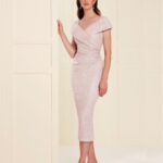 Woman wearing a pale pink sequin midi dress with wrap detailing, a matching light pink hat, and nude heels, posing indoors against cream panels.