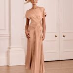 Woman in a peach satin gown with a draped waist and pleated sash, wearing a wide-brim hat, standing in a light-filled paneled room.