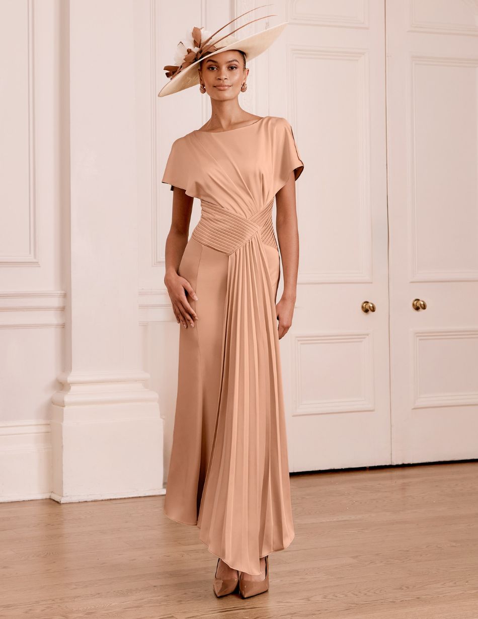 Woman in a peach satin gown with a draped waist and pleated sash, wearing a wide-brim hat, standing in a light-filled paneled room.