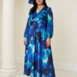 Woman in a blue abstract floral maxi dress with long sleeves, a wrapped waist, and a large blue hat, standing indoors.