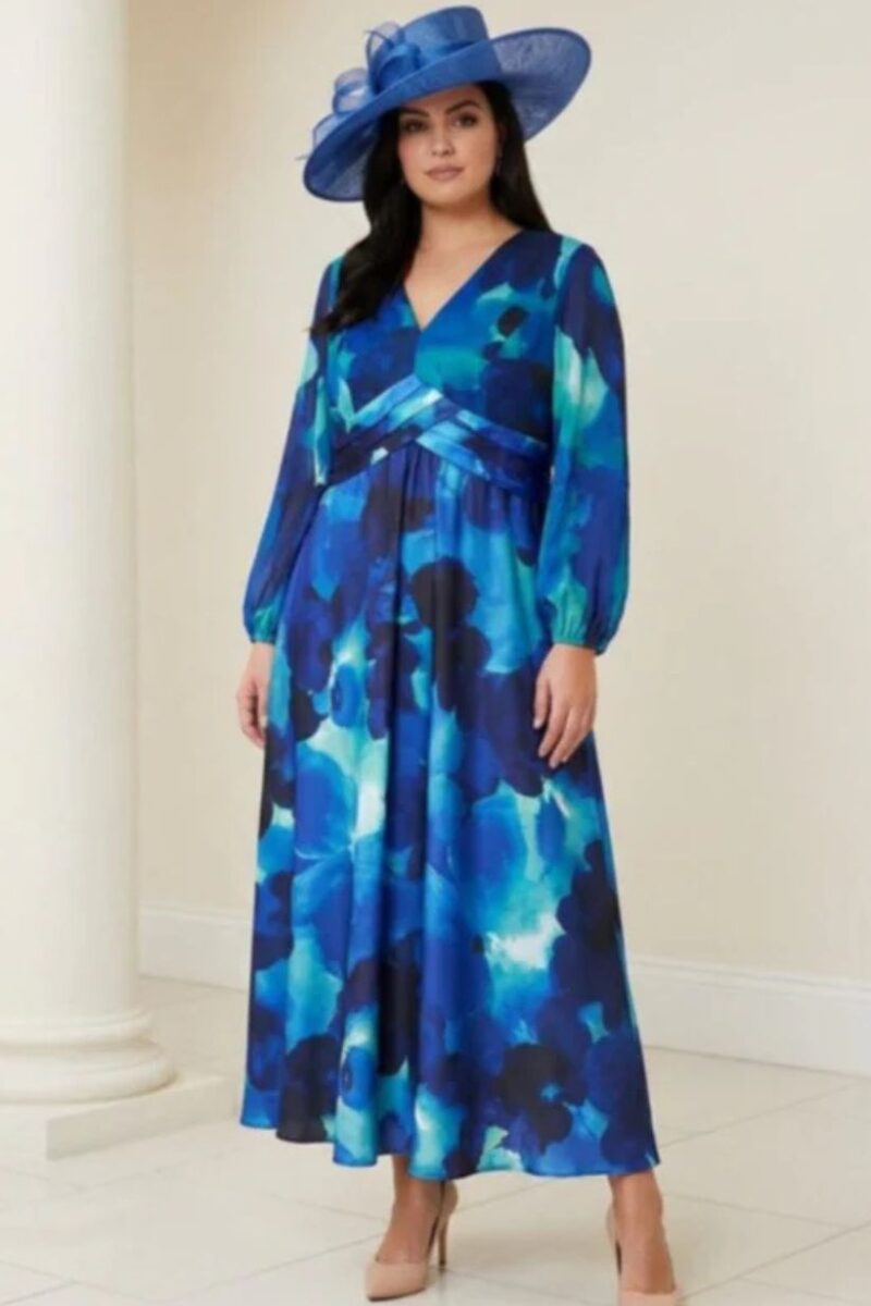 Woman in a blue abstract floral maxi dress with long sleeves, a wrapped waist, and a large blue hat, standing indoors.