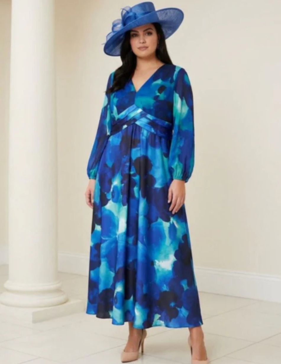 Woman in a blue abstract floral maxi dress with long sleeves, a wrapped waist, and a large blue hat, standing indoors.