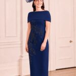 Woman in a navy blue midi dress with floral embroidery and an off-the-shoulder overlay, wearing a large navy fascinator, indoors.