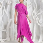 Woman in a bright pink one-shoulder dress with a tied waist and pleated asymmetric hem, standing against white sculptural background.