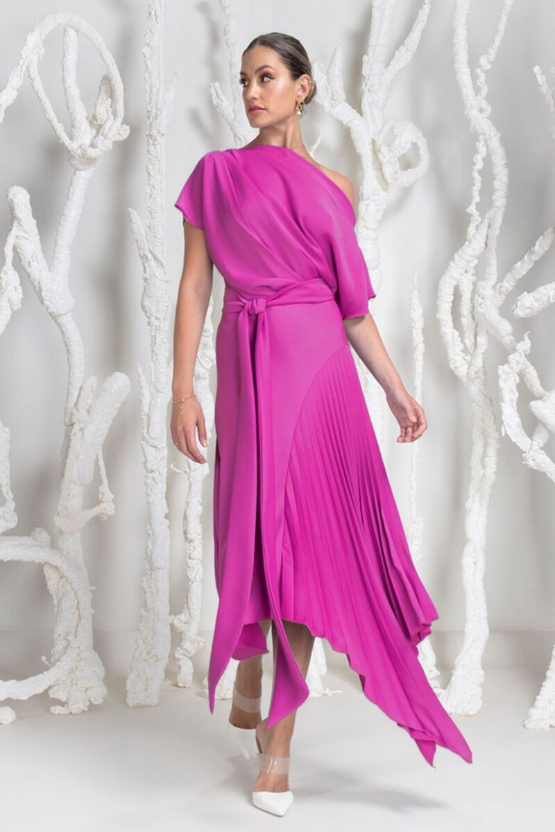 Woman in a bright pink one-shoulder dress with a tied waist and pleated asymmetric hem, standing against white sculptural background.