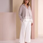 Woman in a light gray oversized sweater and white wide-leg pants posing in a pastel studio setup.
