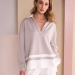 Model wearing an oversized taupe sweater with a white hem and white pants, standing against a pastel backdrop.