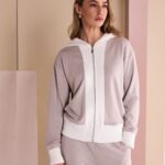 Woman modeling a light gray zip-up hoodie with white cuffs and waistband in a pastel-studio setting