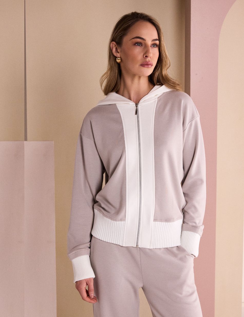 Woman modeling a light gray zip-up hoodie with white cuffs and waistband in a pastel-studio setting