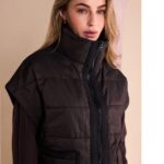 Woman wearing a dark brown quilted puffer vest with a high zip collar, posed indoors.
