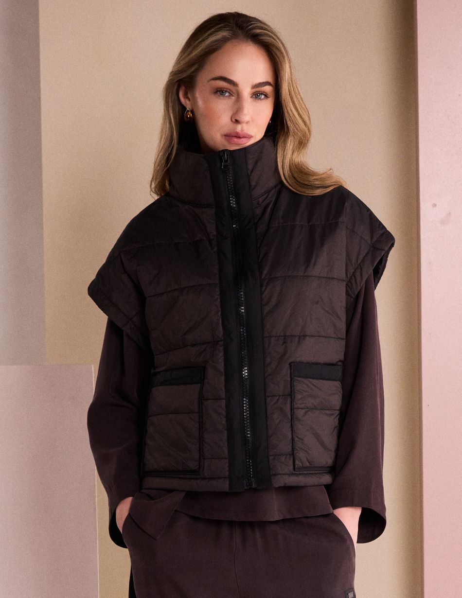 NAW26167 Front Woman wearing a black padded vest over a dark brown outfit, standing with hands in pockets indoors.