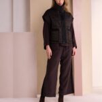 Model wearing a dark brown puffer vest over an oversized top and wide-leg cropped pants, paired with brown ankle boots, standing in a pastel-studio backdrop.