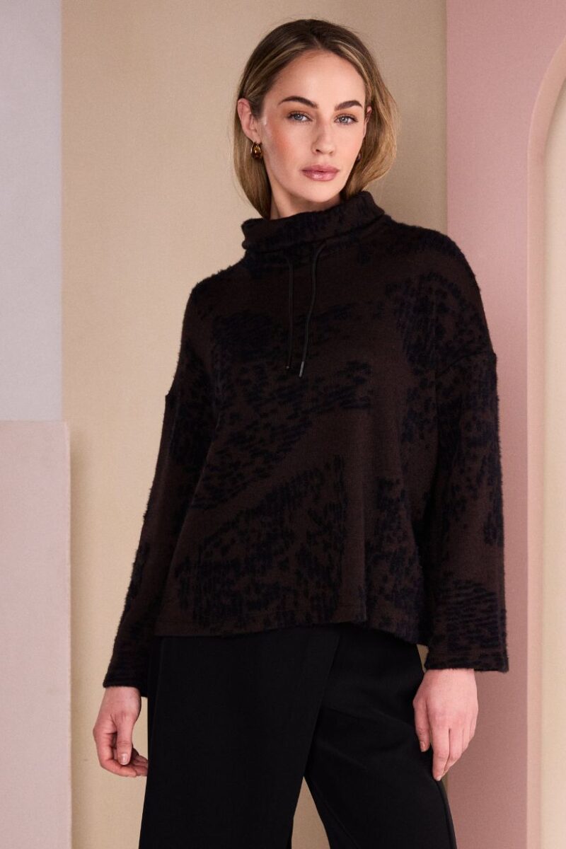 Model wearing a black fuzzy textured turtleneck sweater with drawstrings, standing against pastel walls in a studio setting.