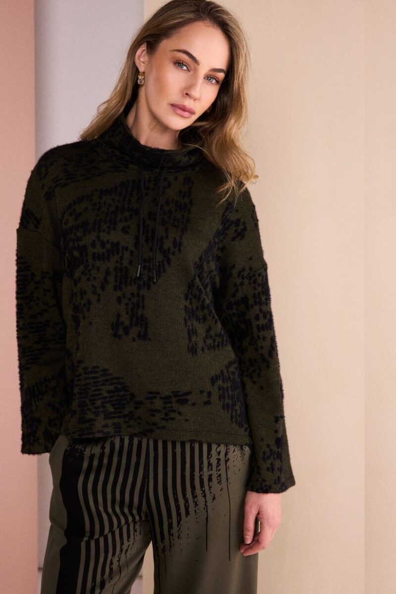 Woman wearing an oversized olive-green knit sweater with a black abstract pattern, standing against a beige wall indoors.