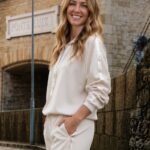 Smiling woman in a cream tracksuit stands on a cobbled street beside a stone building, hands in pockets.