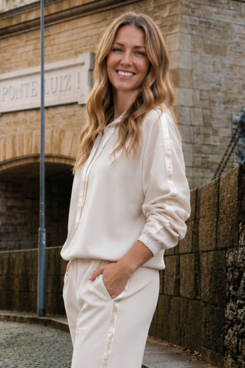 Smiling woman in a cream tracksuit stands on a cobbled street beside a stone building, hands in pockets.