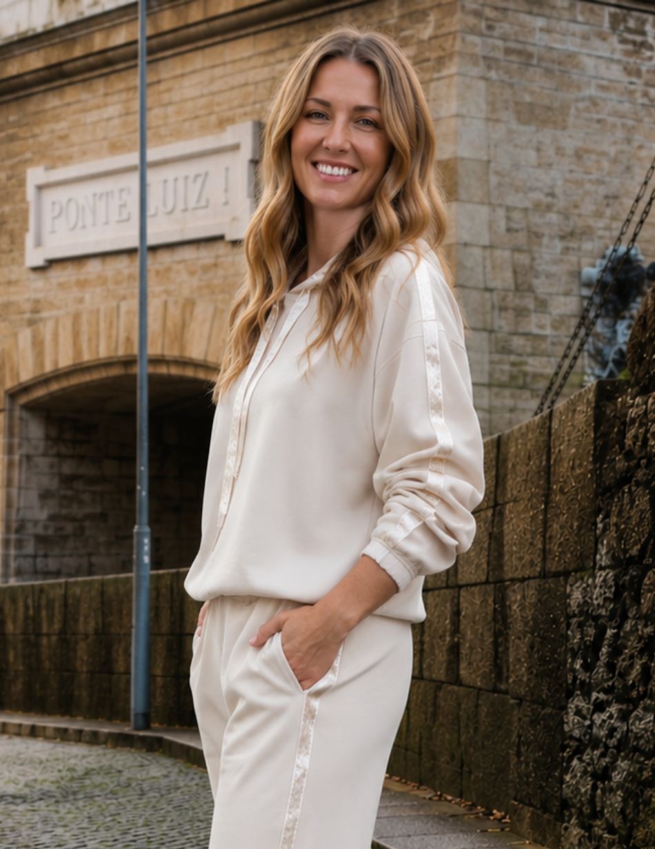 Smiling woman in a cream tracksuit stands on a cobbled street beside a stone building, hands in pockets.